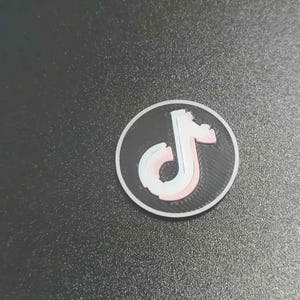 Pu&ograve; includere: Un adesivo rotondo, bianco e nero con il logo di TikTok. Il logo &egrave; una nota musicale stilizzata in bianco, rosa e blu. L'adesivo &egrave; su una superficie grigio scuro.