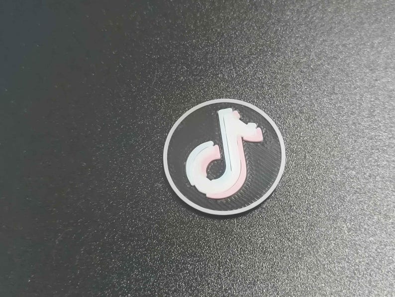 Pu&ograve; includere: Un adesivo rotondo, bianco e nero con il logo di TikTok. Il logo &egrave; una nota musicale stilizzata in bianco, rosa e blu. L'adesivo &egrave; su una superficie grigio scuro.