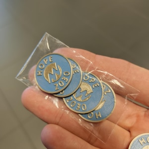 Pu&ograve; includere: Una collezione di spille rotonde blu e oro, ognuna con la scritta "HOPE" e il numero "2030". Le spille sono confezionate in un sacchetto di plastica trasparente. Le spille hanno un emblema dorato al centro.