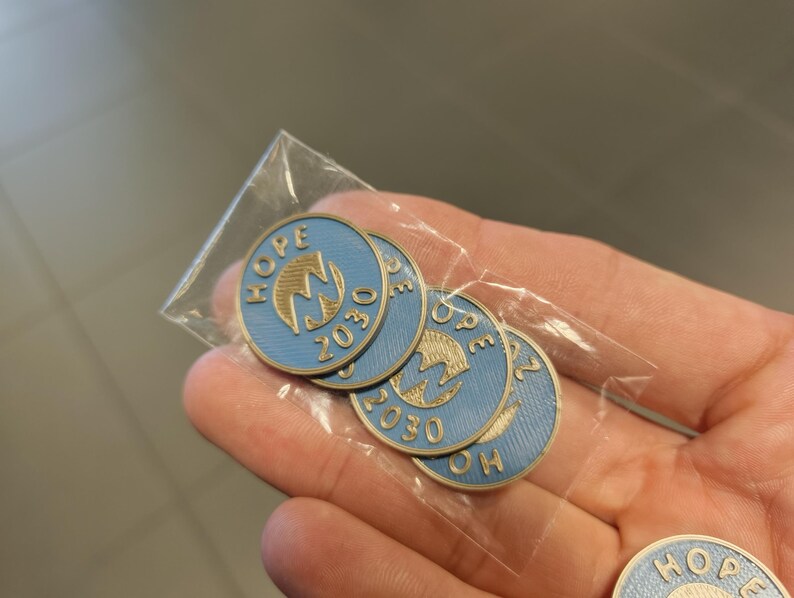 Pu&ograve; includere: Una collezione di spille rotonde blu e oro, ognuna con la scritta "HOPE" e il numero "2030". Le spille sono confezionate in un sacchetto di plastica trasparente. Le spille hanno un emblema dorato al centro.