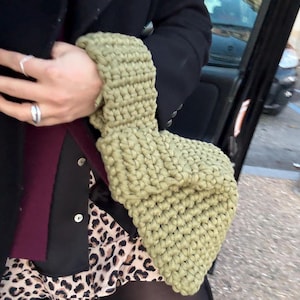 Può includere: Una borsa a mano verde chiaro a maglia grossa. La borsa è realizzata con un filato spesso e strutturato e ha una forma rettangolare. La borsa è tenuta da una persona che indossa un cappotto nero e una gonna con stampa leopardata.