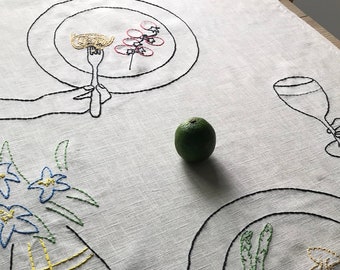 Può includere: Tovaglia di lino bianco ricamata con una mano che tiene una forchetta sopra un piatto di pasta, un bicchiere di vino, un vaso di fiori blu, un lime e un piatto di asparagi.