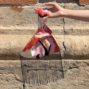 Op de afbeelding: Een handtas met patroon in een driehoekige vorm en een zwarte franje. De tas heeft een handvat en een ontwerp met rode, roze en zwarte abstracte vormen. De tas wordt door een hand omhoog gehouden.