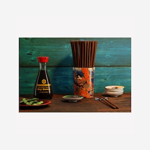 May include: A wooden table with a blue and green wooden background. A bottle of Kikkoman soy sauce, a small dish of soy sauce, a plate of edamame, a set of chopsticks in a ceramic holder with a Japanese design, and a stack of small white plates.