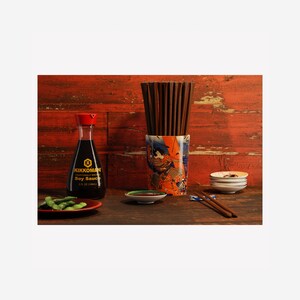 Puede incluir: Una mesa puesta con una botella de salsa de soja Kikkoman, un pequeño plato de salsa de soja, un plato de edamame, un porta-palillos de cerámica con un diseño japonés y palillos. El fondo es una pared de madera roja.