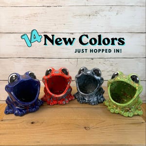 May include: Four ceramic frog-shaped containers in blue, red, grey, and green with speckled finishes. The text above reads 14 New Colors JUST HOPPED IN! Each frog has a wide open mouth, large black eyes, and a glossy glazed surface texture.