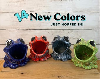 Könnte beinhalten: Vier Keramikbehälter in Froschform in Blau, Rot, Grau und Grün mit gesprenkelten Oberflächen. Der Text lautet 14 New Colors JUST HOPPED IN! Jeder Frosch hat ein weit geöffnetes Maul, große schwarze Augen und eine glänzende Glasur.