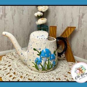 May include: A white ceramic watering can with blue splatter paint and a black handle. The can is decorated with blue mushroom illustrations and green grass. The watering can is sitting on a white crocheted doily.