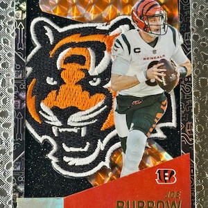 May include: A sports trading card featuring a football player in a white and black uniform, holding a football. The card has a black and orange tiger logo and the name "BURROW" in gold lettering. The card is framed with a silver border.