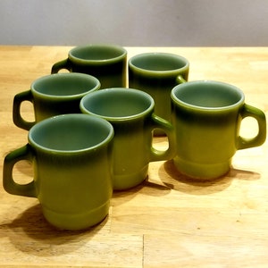 Pode incluir: Seis canecas de vidro verde com interior branco. As canecas têm uma forma arredondada e uma alça na lateral.