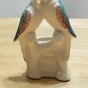 以下が含まれることがあります： 枝に止まっている2羽の鳥の形をした白い陶器製のプランター。鳥は緑、青、ピンクのアクセントで描かれています。