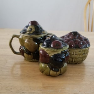 Pode incluir: Um conjunto de três potes de cozinha de cerâmica com desenhos de frutas e bagas. Os potes são verdes, marrons e pretos com um visual rústico.