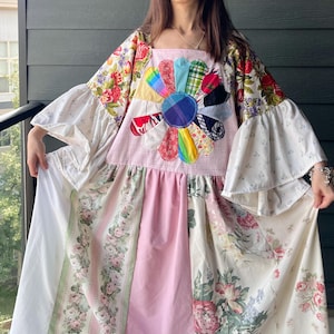 May include: A floor-length patchwork dress featuring a large central flower appliqué made of various colourful fabric petals. The garment includes wide bell sleeves, a square neckline, and a skirt composed of panels with floral and solid pink patterns.