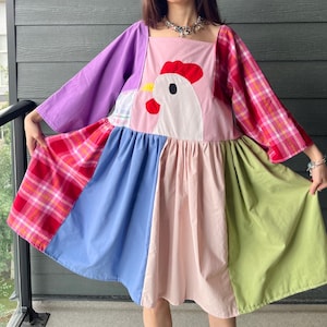 May include: A colour-blocked dress featuring a large white chicken appliqué with a red comb and wattle on a pink bodice. The garment includes long sleeves in purple and pink plaid, with a gathered skirt panelled in red plaid, blue, pink, and light green fabric.