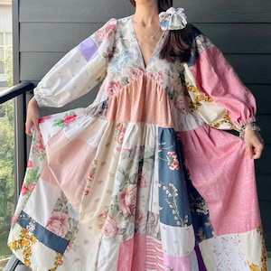 Pode incluir: Um vestido patchwork de manga comprida com decote em V e um elástico de cabelo a combinar. A peça é construída a partir de vários painéis de tecido em tons de rosa, branco, azul e lavanda, decorados com padrões florais e texturas lisas.