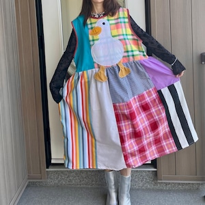 May include: A sleeveless patchwork dress featuring a large white duck appliqué with an orange beak and feet. The dress is constructed from various fabric panels including colorful vertical stripes, a yellow and green plaid, blue gingham, and pink plaid.