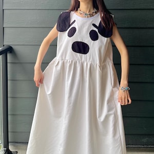 May include: A white sleeveless dress featuring a large black graphic of a dog face on the chest, complete with two black fabric ears attached at the shoulders. The dress has a round neckline and a gathered waistline that flows into a long skirt.