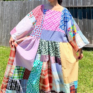 May include: A patchwork dress featuring a variety of fabric patterns and colors, including gingham, floral, and solid patches. The dress has a square neckline and wide sleeves. The dress is knee-length and appears to be handmade.