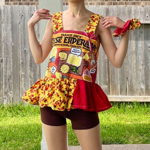 May include: A handmade, sleeveless top and matching scrunchie. The top is made from a Trader Joe's "ESE EXPERA" cheese grits box, with a red and yellow chili pepper print on the skirt. The scrunchie has the same chili pepper print.
