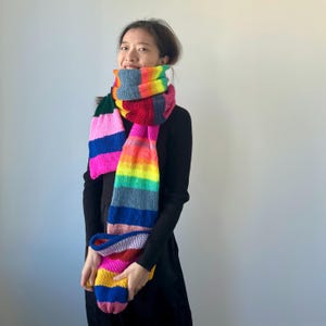 May include: A long, colorful knitted scarf with a rainbow pattern. The scarf features stripes of pink, blue, green, yellow, orange, and red. The scarf is wrapped around the neck and held in the hands.