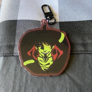 May include: A black and green anime character keychain with a red and black background. The character has a green face and is wearing a black coat with a red collar.