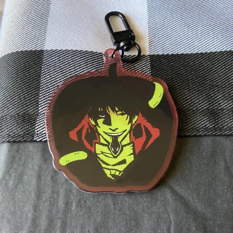 May include: A black and green anime character keychain with a red and black background. The character has a green face and is wearing a black coat with a red collar.