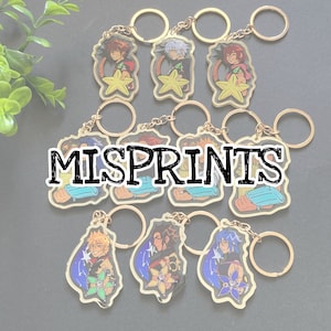 May include: A collection of gold-colored keychains featuring various anime-style character designs. Each keychain has a clear acrylic charm with a character illustration and a gold-colored keyring. The text "MISPRINTS" is overlaid on the image.