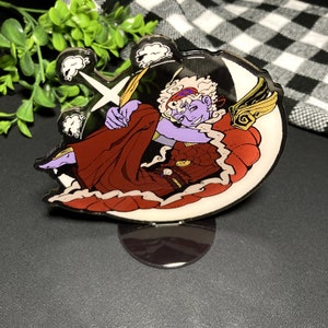 Peut inclure: Un personnage violet et rouge avec des ailes est assis sur une lune croissante avec des nuages et des éclairs derrière lui. Le personnage porte une couronne et une robe rouge. L'image est imprimée sur un support en acrylique transparent.