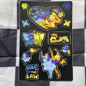 May include: A black sticker sheet featuring multiple illustrations of two characters, one purple and one yellow, in various poses. The designs include a heart-shaped frame, a cowboy with a gun, and the text "LOVE IS THE LAW" in blue and yellow.