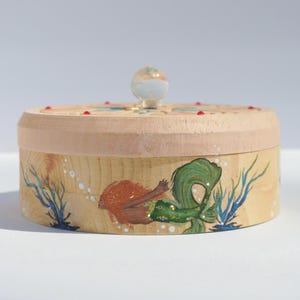 May include: A wooden box with a clear glass knob on the lid. The box is painted with a mermaid swimming in the ocean with seaweed and bubbles.