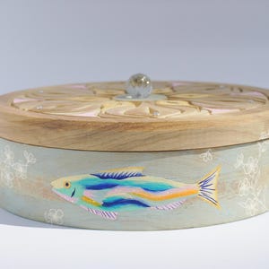 May include: A round wooden box with a light blue painted exterior. The lid is decorated with a carved floral design and a glass knob. The box features a painted image of a colorful fish with white floral accents.