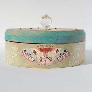 May include: A round wooden box with a teal blue lid and a painted butterfly design. The box has a clear glass knob on the lid.