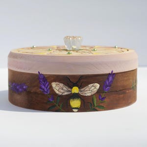 May include: A wooden box with a pink lid and a painted design of a bee and lavender flowers. The box has a clear glass knob on the lid.