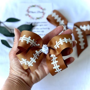 May include: A brown and white football-themed hair bow. The bow is made of brown ribbon with white stitching details, resembling a football. A smaller white bow is in the center. Additional bows are visible in the background.