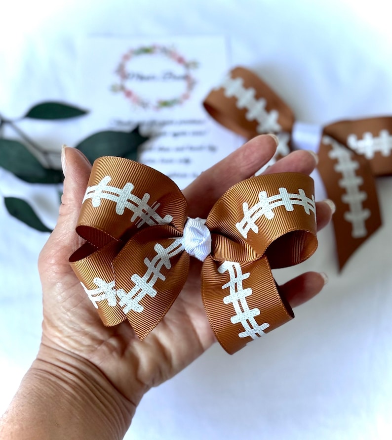 May include: A brown and white football-themed hair bow. The bow is made of brown ribbon with white stitching details, resembling a football. A smaller white bow is in the center. Additional bows are visible in the background.