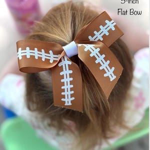 May include: A brown and white football-themed flat bow, approximately 5 inches in size. The bow features a football design with white stitching details. The bow is secured with a white band.