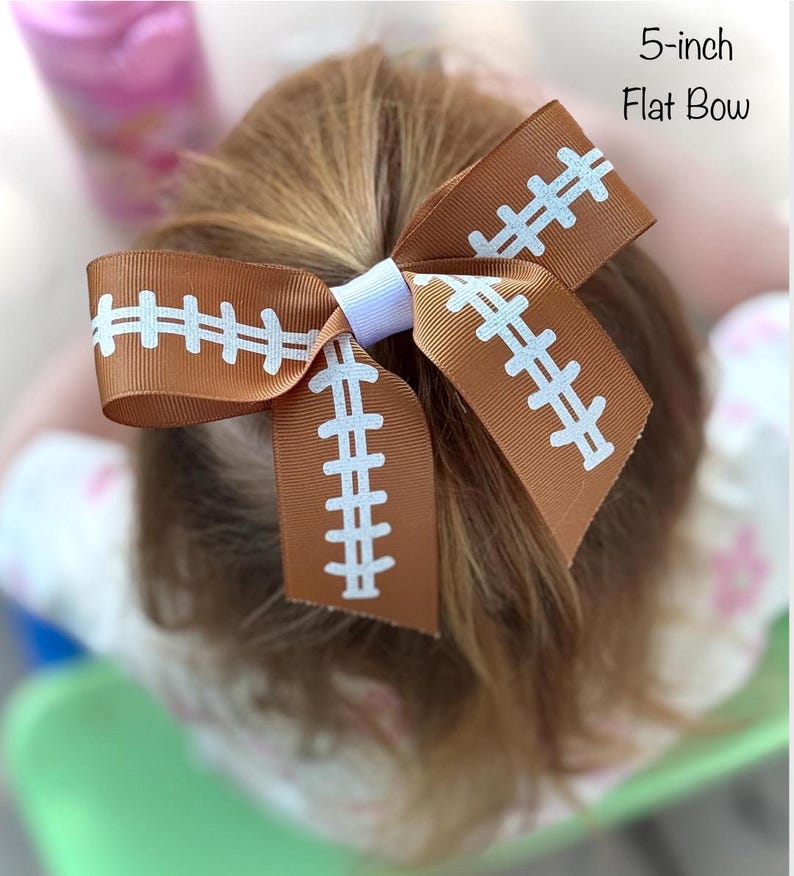 May include: A brown and white football-themed flat bow, approximately 5 inches in size. The bow features a football design with white stitching details. The bow is secured with a white band.