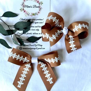 May include: Two brown and white football-themed hair bows. The bows are made of ribbon and feature a football design with white stitching. The bows are arranged on a white surface with a business card and greenery.