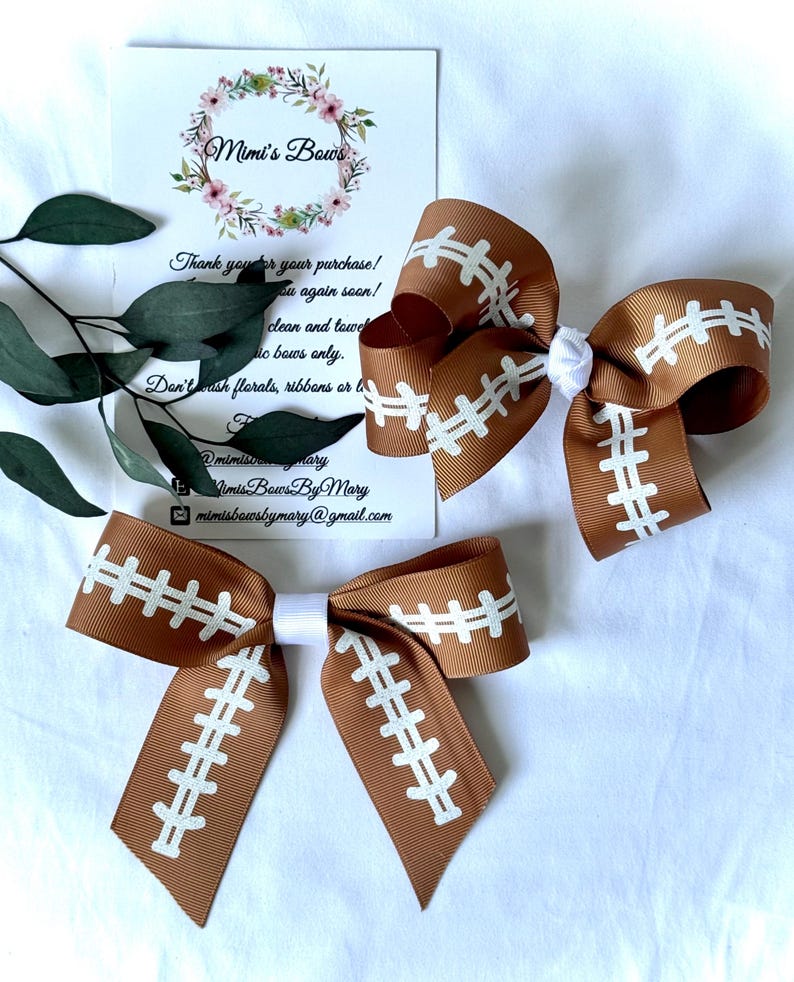 May include: Two brown and white football-themed hair bows. The bows are made of ribbon and feature a football design with white stitching. The bows are arranged on a white surface with a business card and greenery.
