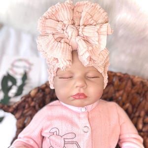 May include: A baby doll wearing a light pink turban-style headband with a large bow. The doll is also wearing a light pink onesie with a bunny graphic. The doll is in a woven basket.
