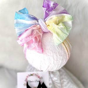May include: A large, multi-colored fabric hair bow featuring a crinkled texture in shades of blue, purple, yellow, green, and pink. The bow is attached to a white lace headband. A small white card with a floral wreath logo is visible below.