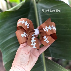 May include: A brown football-themed bow, 5 inches wide, with white stitching detail. The bow is made of ribbon and has a white center knot. The text "5-Inch Wide Bow" is visible in the image.