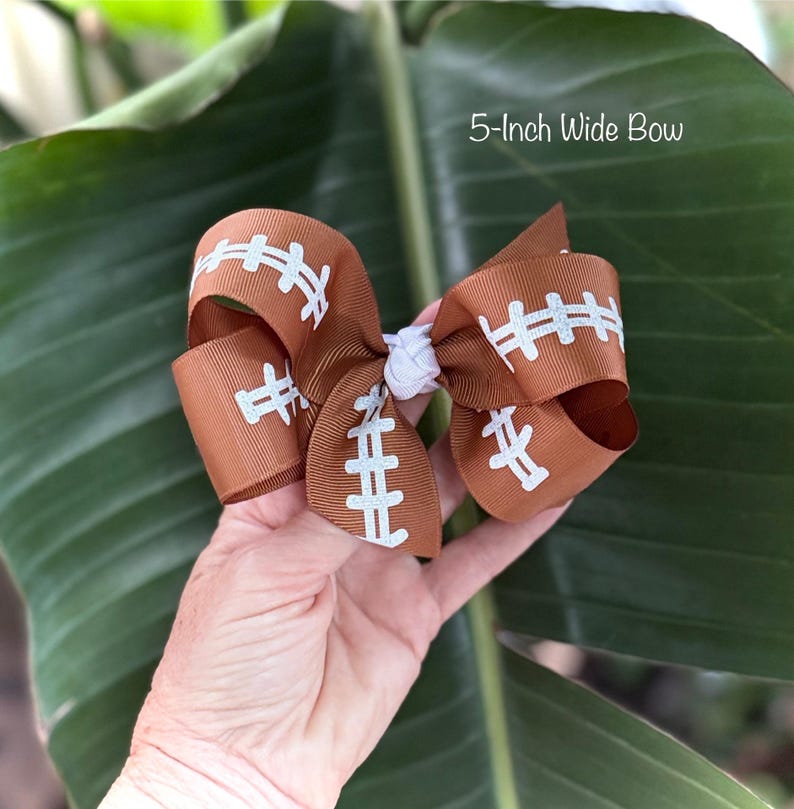 May include: A brown football-themed bow, 5 inches wide, with white stitching detail. The bow is made of ribbon and has a white center knot. The text "5-Inch Wide Bow" is visible in the image.