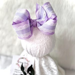 May include: A large, lavender hair bow with white lace trim. The bow is made of a wide ribbon and is tied in a classic bow shape. The bow is displayed on a white, textured surface. A card with text is visible.