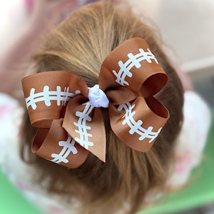 May include: Brown hair bow with a football design. The bow is made of brown ribbon with white stitching details, resembling a football. A small white flower is in the center of the bow.
