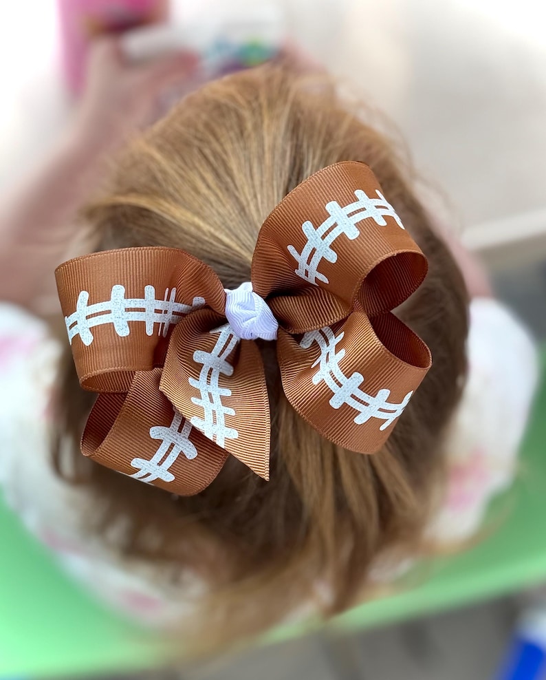 May include: Brown hair bow with a football design. The bow is made of brown ribbon with white stitching details, resembling a football. A small white flower is in the center of the bow.