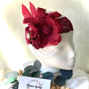 May include: A burgundy headband with a lace trim and a large burgundy flower with a pearl accent. The headband is decorated with a small cluster of burgundy flowers and a single red feather.