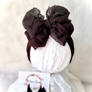 May include: A dark brown baby headband with a large, layered bow. The bow is made of a textured fabric and is attached to a thin, matching headband. The headband is displayed on a white, lace-covered form.