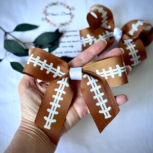May include: Brown and white football-themed hair bows. The bows are made of a brown ribbon with white stitching details, resembling a football. A white ribbon is used to tie the bow together. The bows are held in a hand.