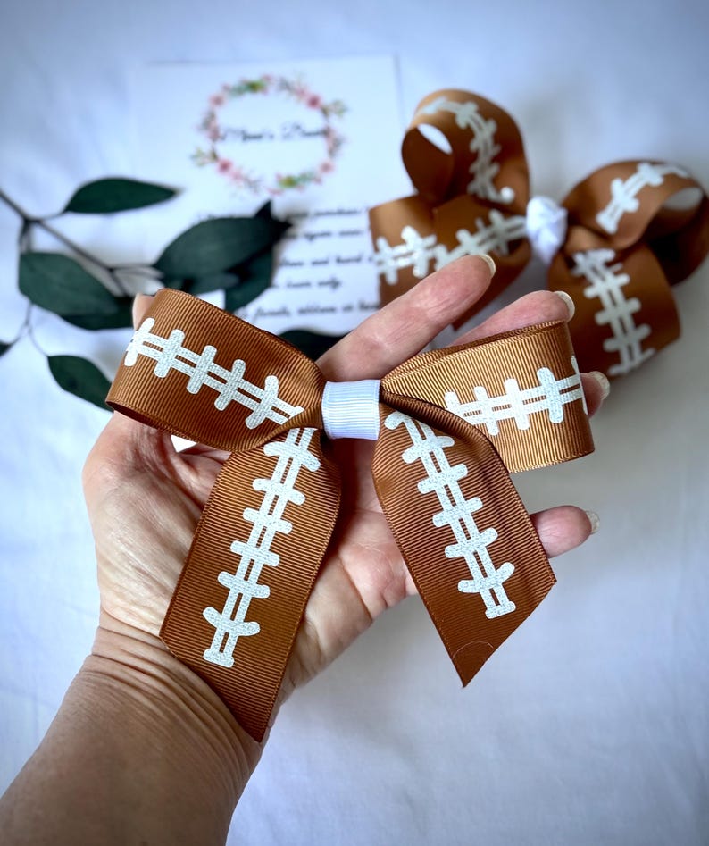 May include: Brown and white football-themed hair bows. The bows are made of a brown ribbon with white stitching details, resembling a football. A white ribbon is used to tie the bow together. The bows are held in a hand.